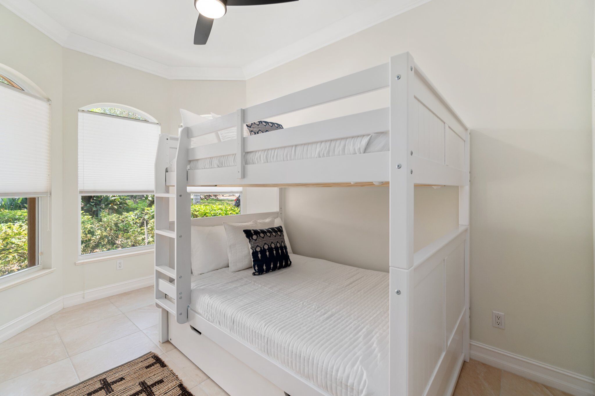 A bunk bed room with white bedding, a ceiling fan, and large windows with blinds.