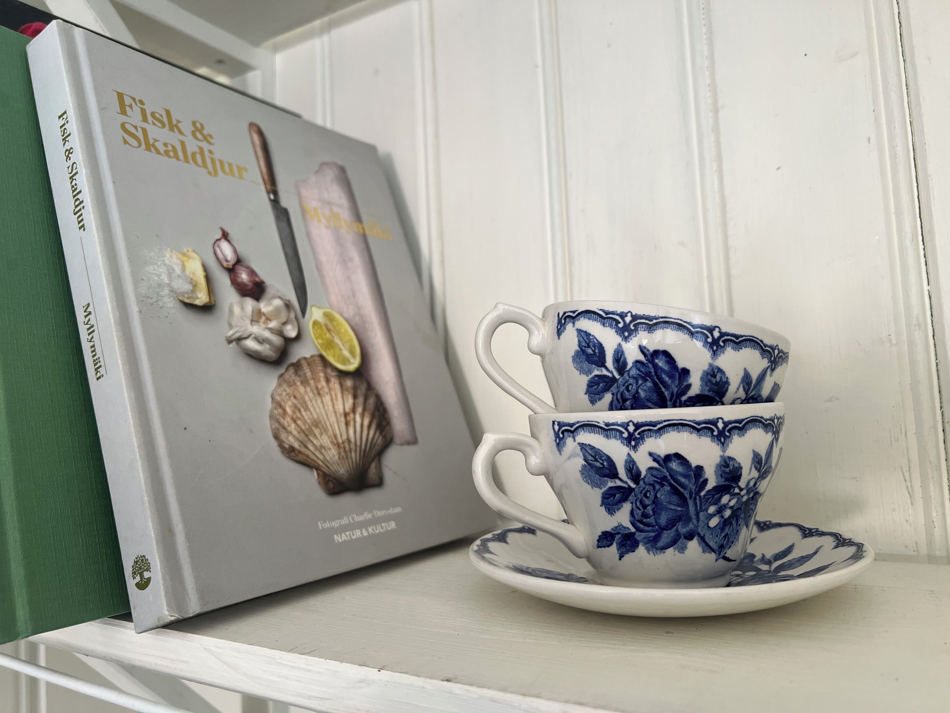 Un libro intitolato 'Fisk & Skaldjur' e una tazza blu con decorazioni floreali su uno scaffale bianco.