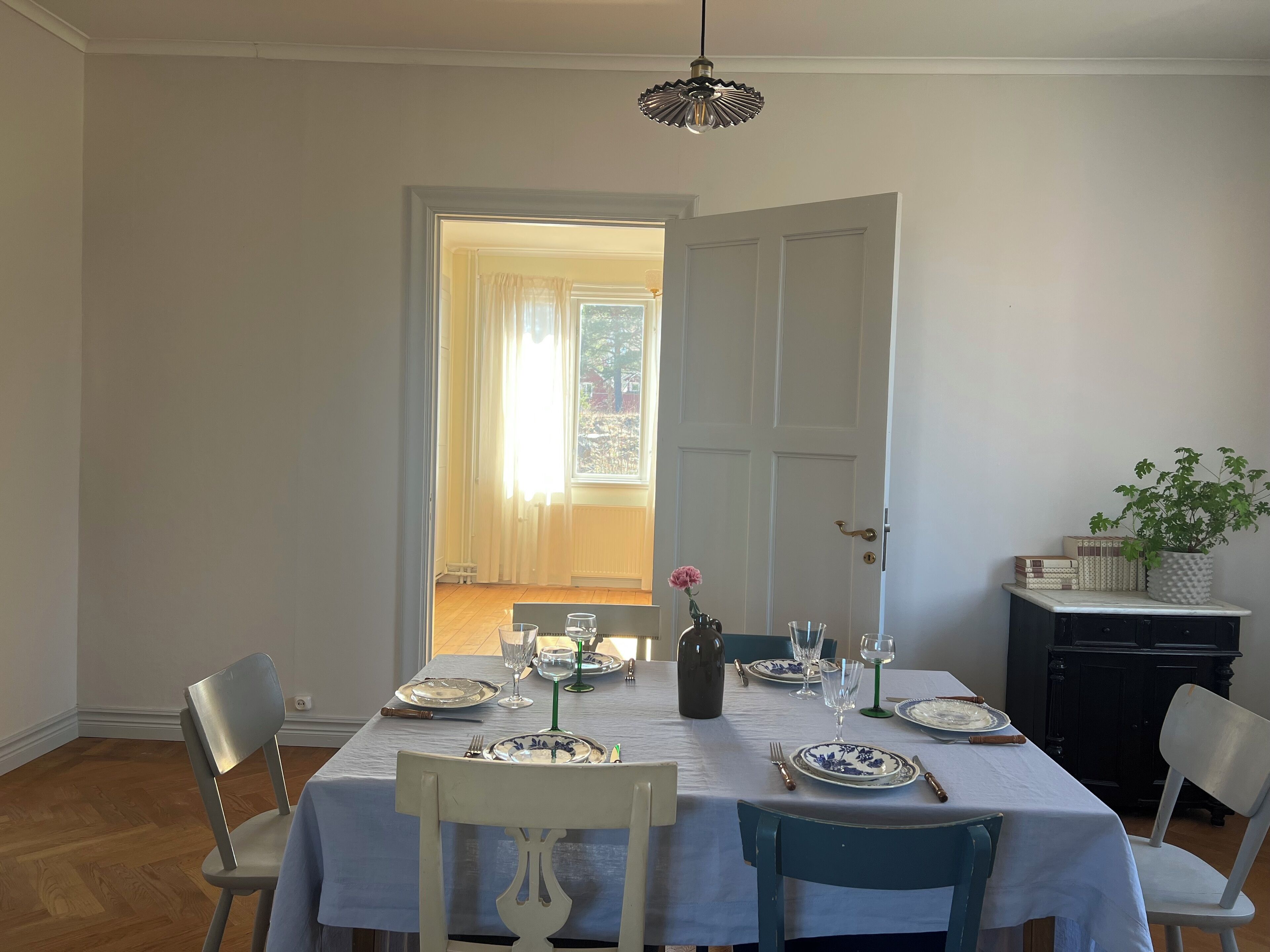 Ein Esszimmer mit einem für sechs Personen gedeckten Tisch, Stühlen, einem Blumentopf und einer Tür zu einem hellen Raum.