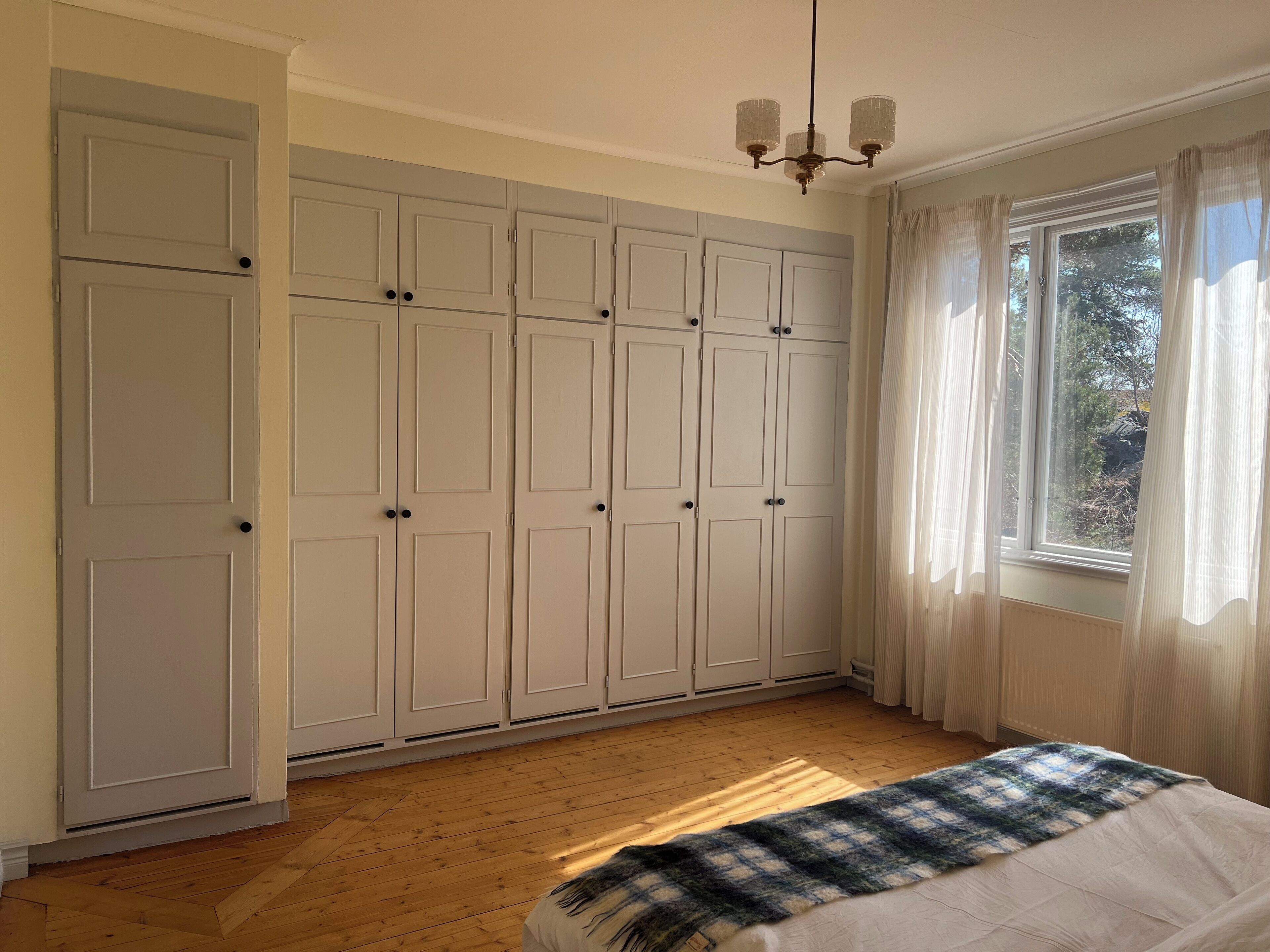 A bedroom with a large wardrobe, a bed with a blue and white blanket, and a window with sheer curtains.