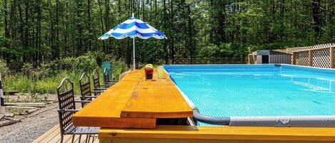 A swimming pool with a wooden deck, a blue and white striped umbrella, and a forested background.