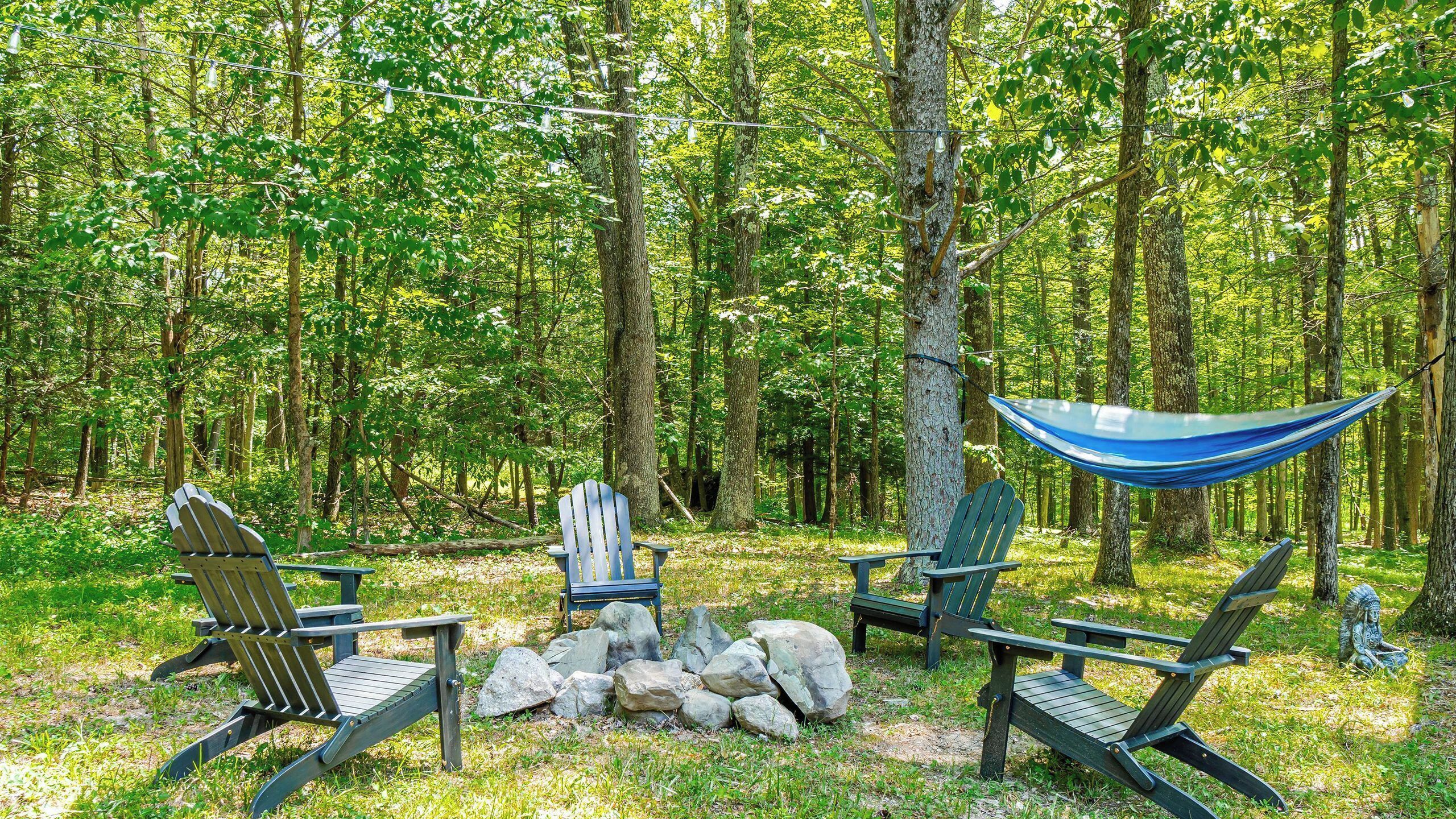 Un cadre forestier avec des espaces de détente en plein air, comprenant des fauteuils Adirondack et un hamac, le tout entouré d’arbres.