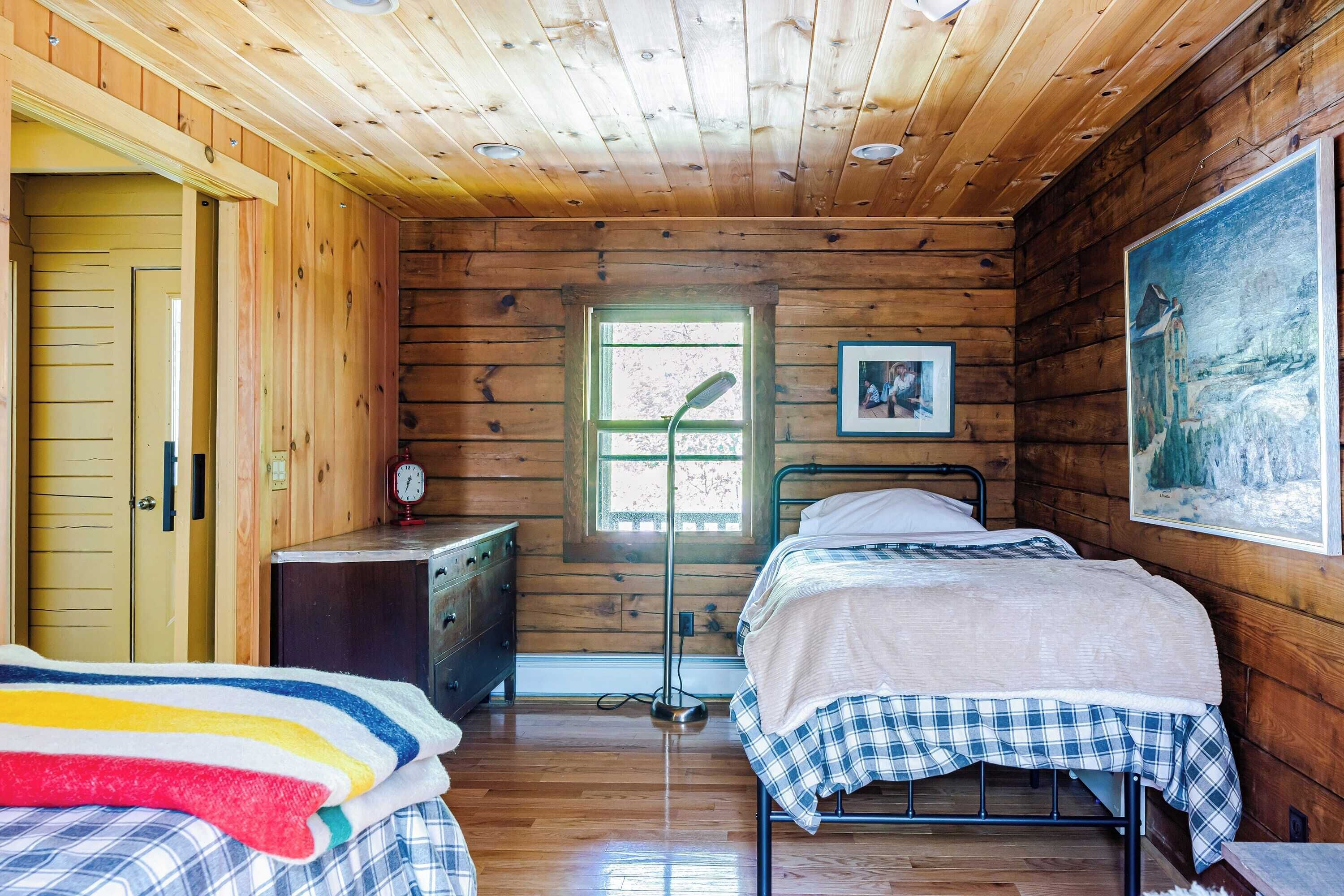 A cozy cabin-style bedroom with wooden walls and ceiling, two beds with plaid bedding, a dresser, a bedside table with a lamp, and a painting on the wall.
