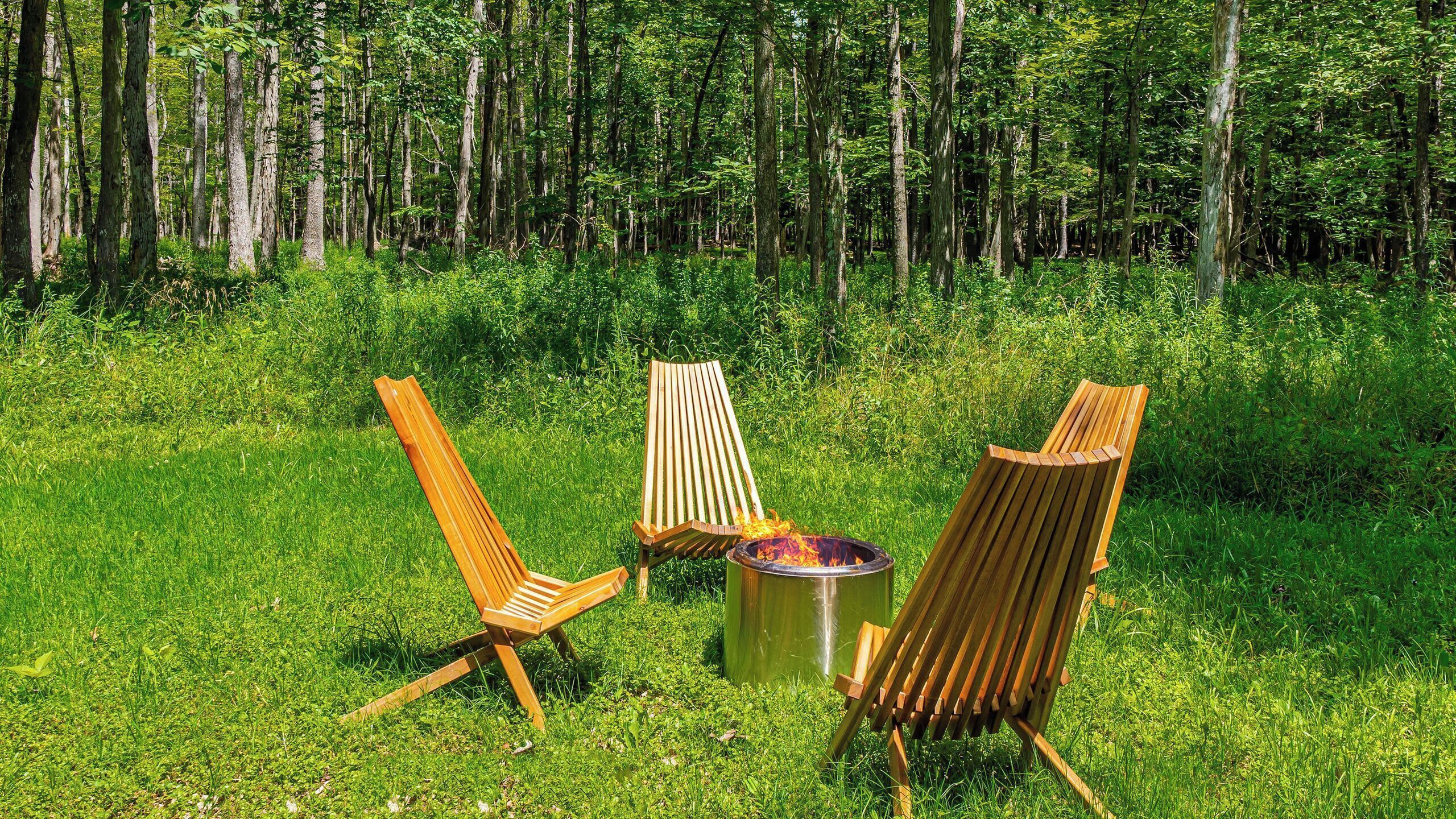 A forested area with three wooden chairs and a fire pit.