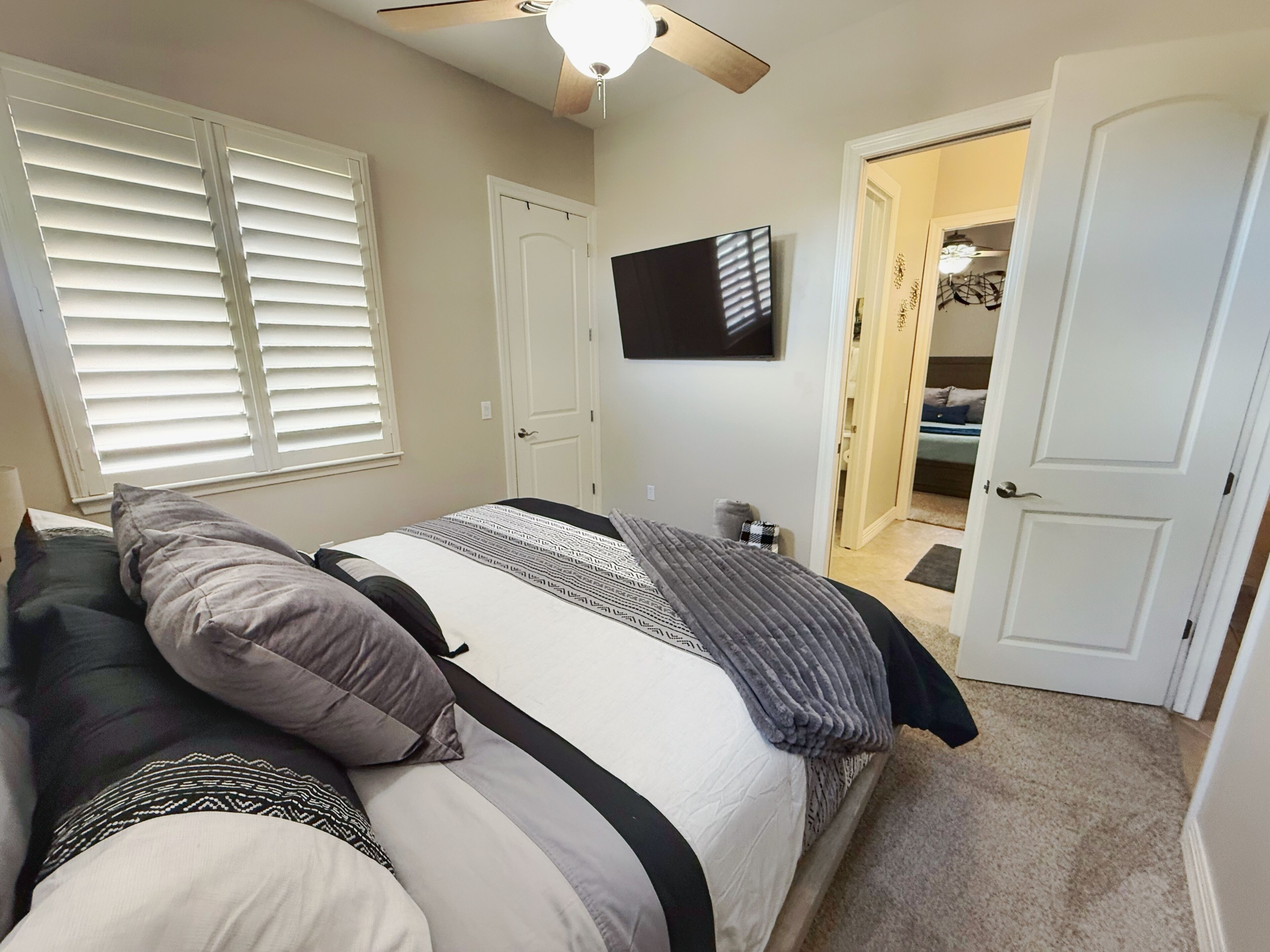 A bedroom with a bed, a ceiling fan, a TV mounted on the wall, and a door leading to another room.
