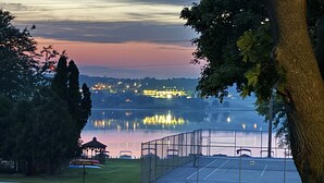 Un parque con un quiosco, una cancha de tenis cercada y un lago que refleja luces.