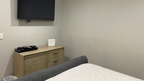 Un dormitorio con una cama, un aparador de madera, un televisor de pantalla plana montado en la pared y una mesita de noche con una lámpara.