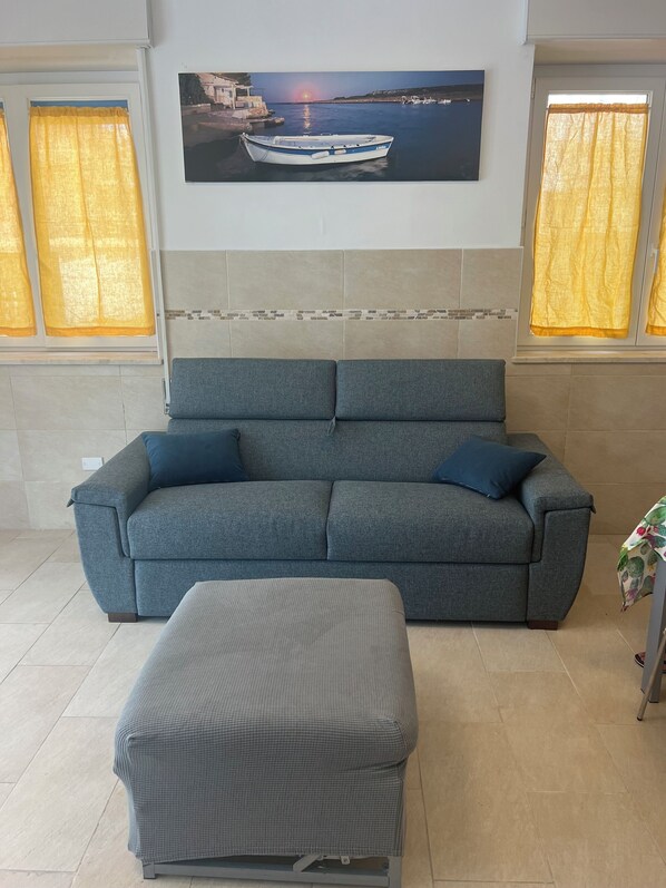 A living room with a grey sofa, a matching ottoman, and a painting of a boat on the wall. - Marzamemi Beach Sun Sea vacation home (Marzamemi)