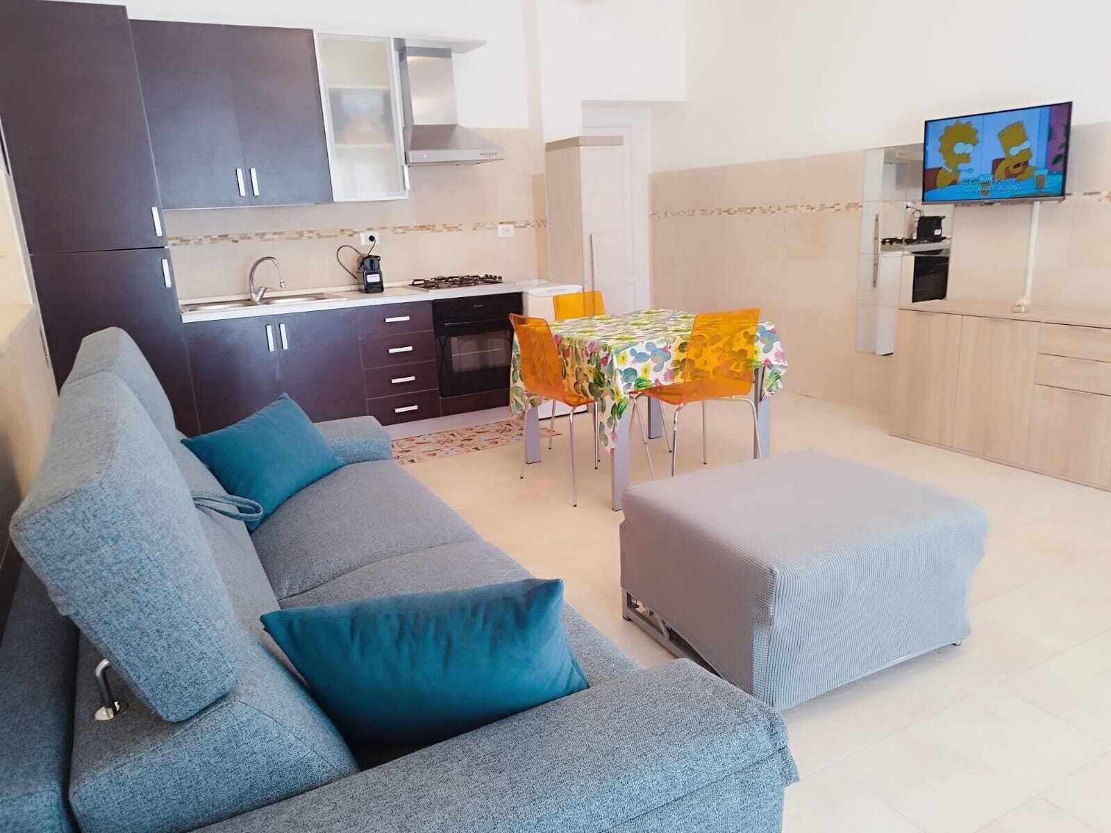 Un soggiorno moderno con un divano grigio, un tavolo da pranzo con sedie, un angolo cottura e una TV a parete.