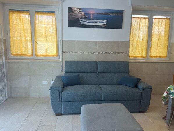 A living room with a grey sofa, a small table with a floral tablecloth, and a painting of a boat on the wall. - Marzamemi Beach Sun Sea vacation home (Marzamemi)