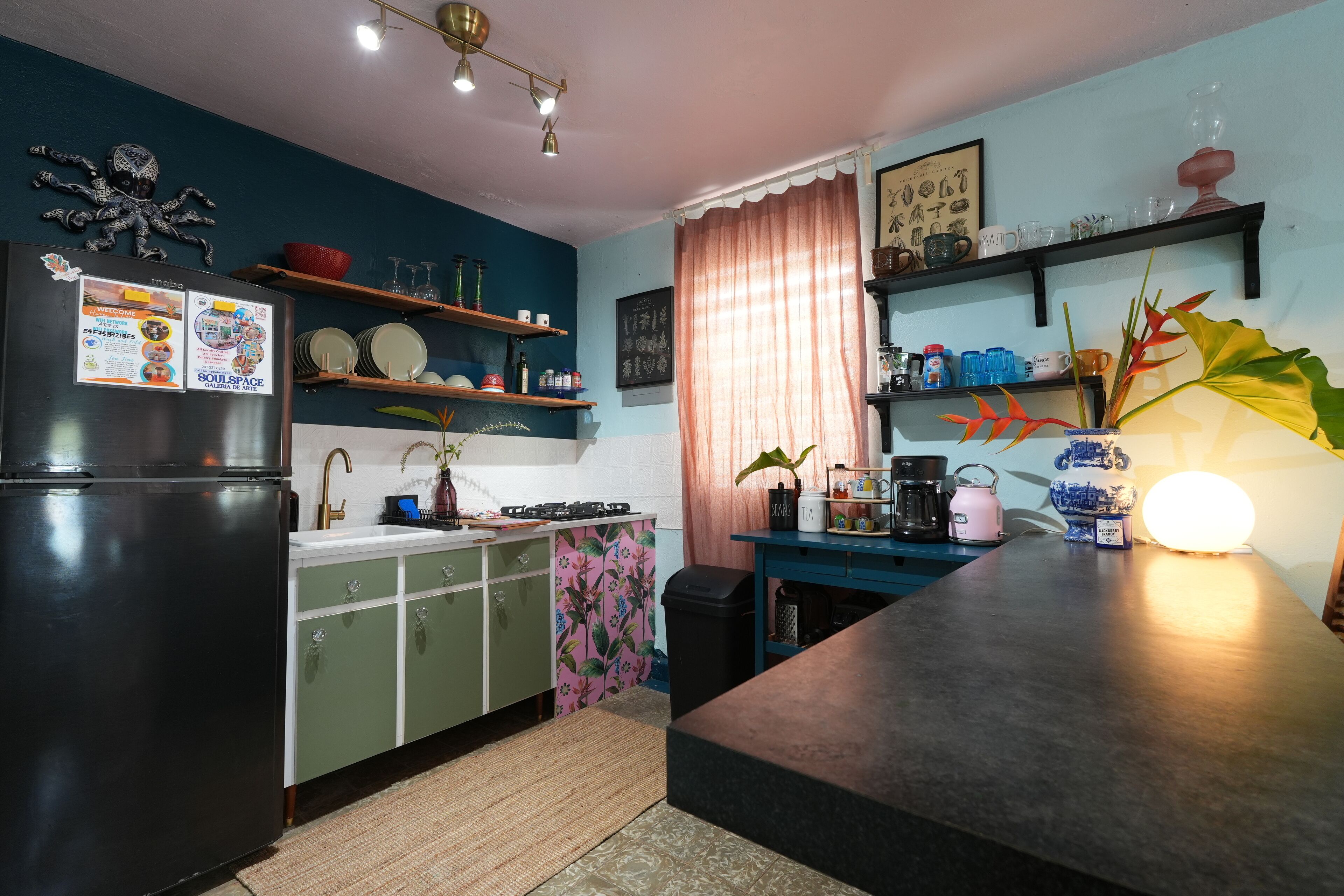 A kitchen with a black refrigerator, green cabinets, a pink floral backsplash, and a blue countertop.