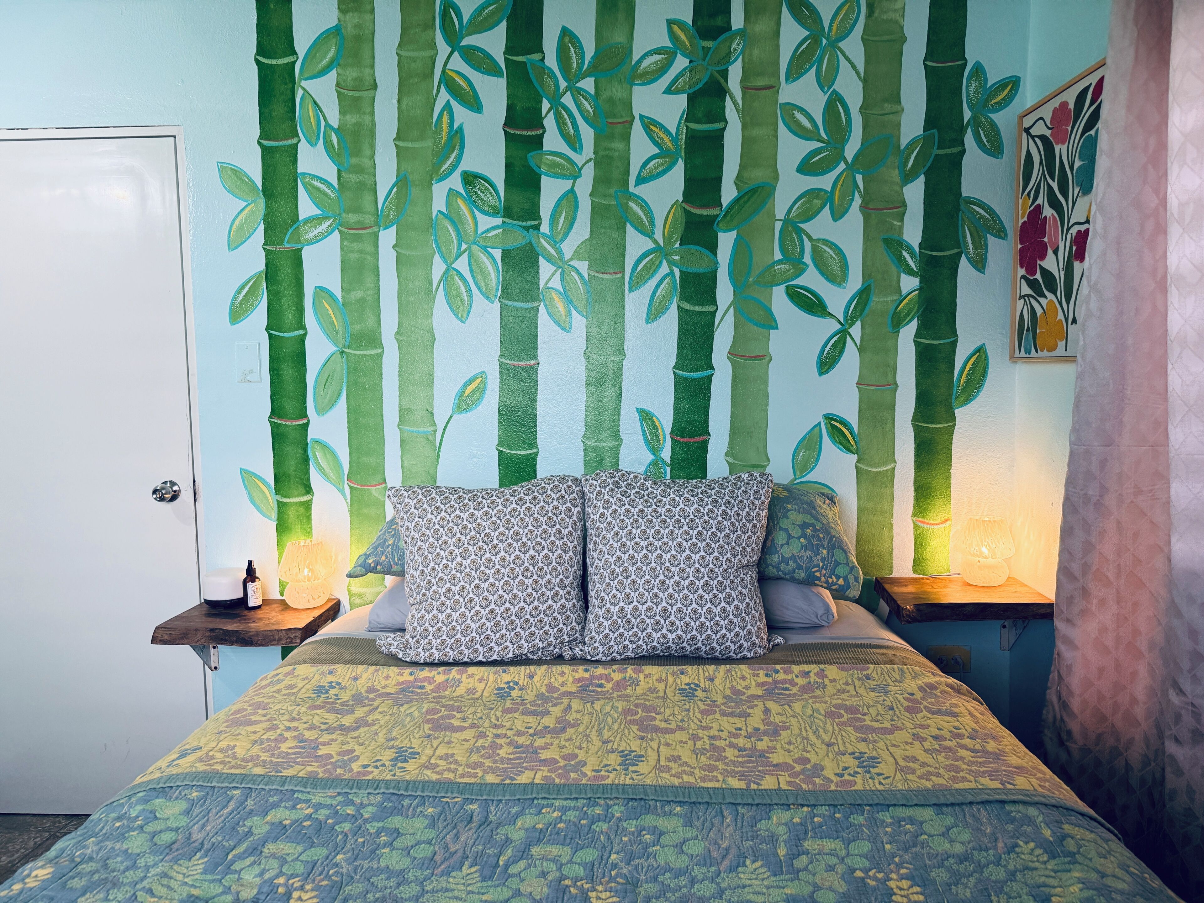 Un dormitorio con una cama, dos almohadas estampadas, una mesita de noche con una lámpara y una pared con motivos de hojas de bambú.