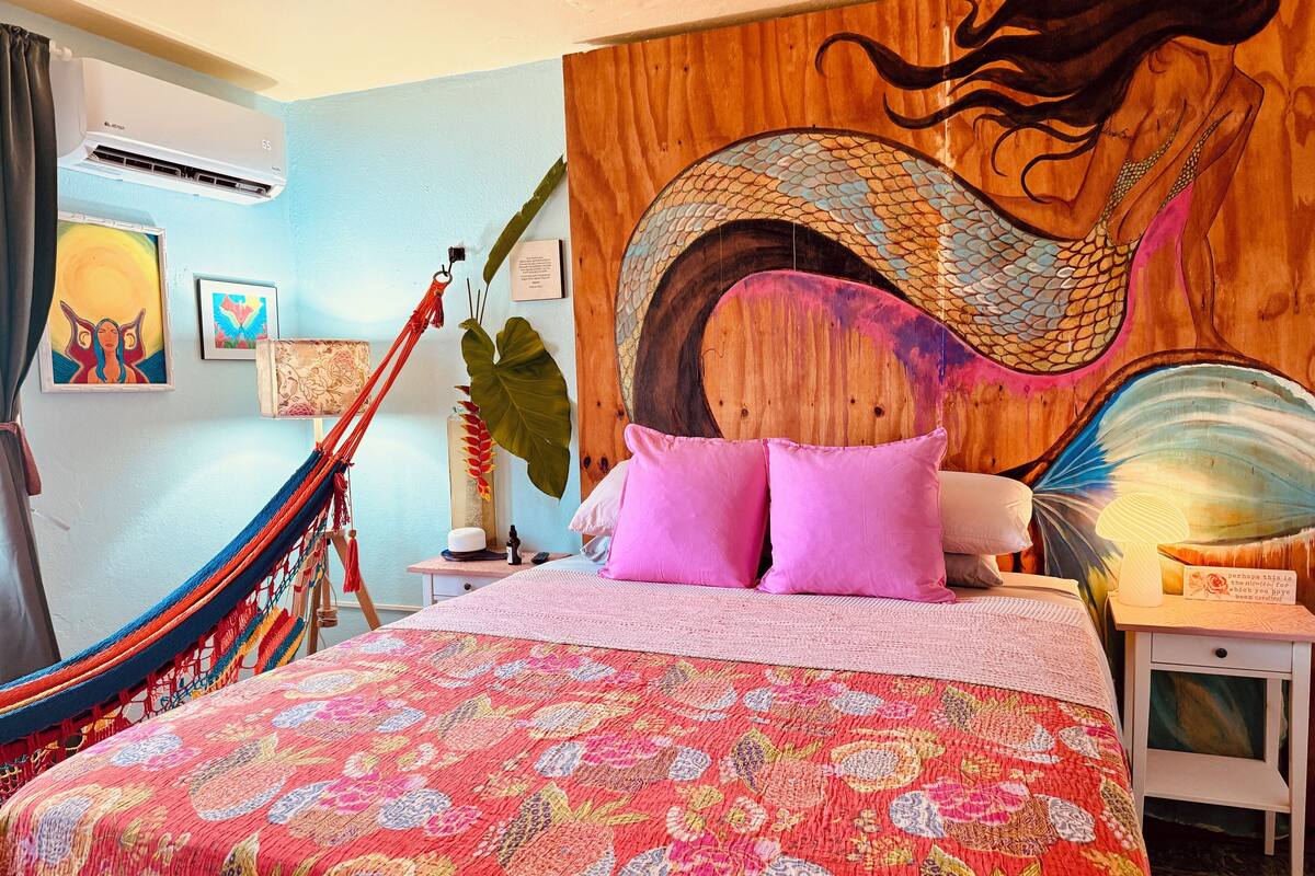 A bedroom with a hammock, a large mural, and a bed with pink pillows.