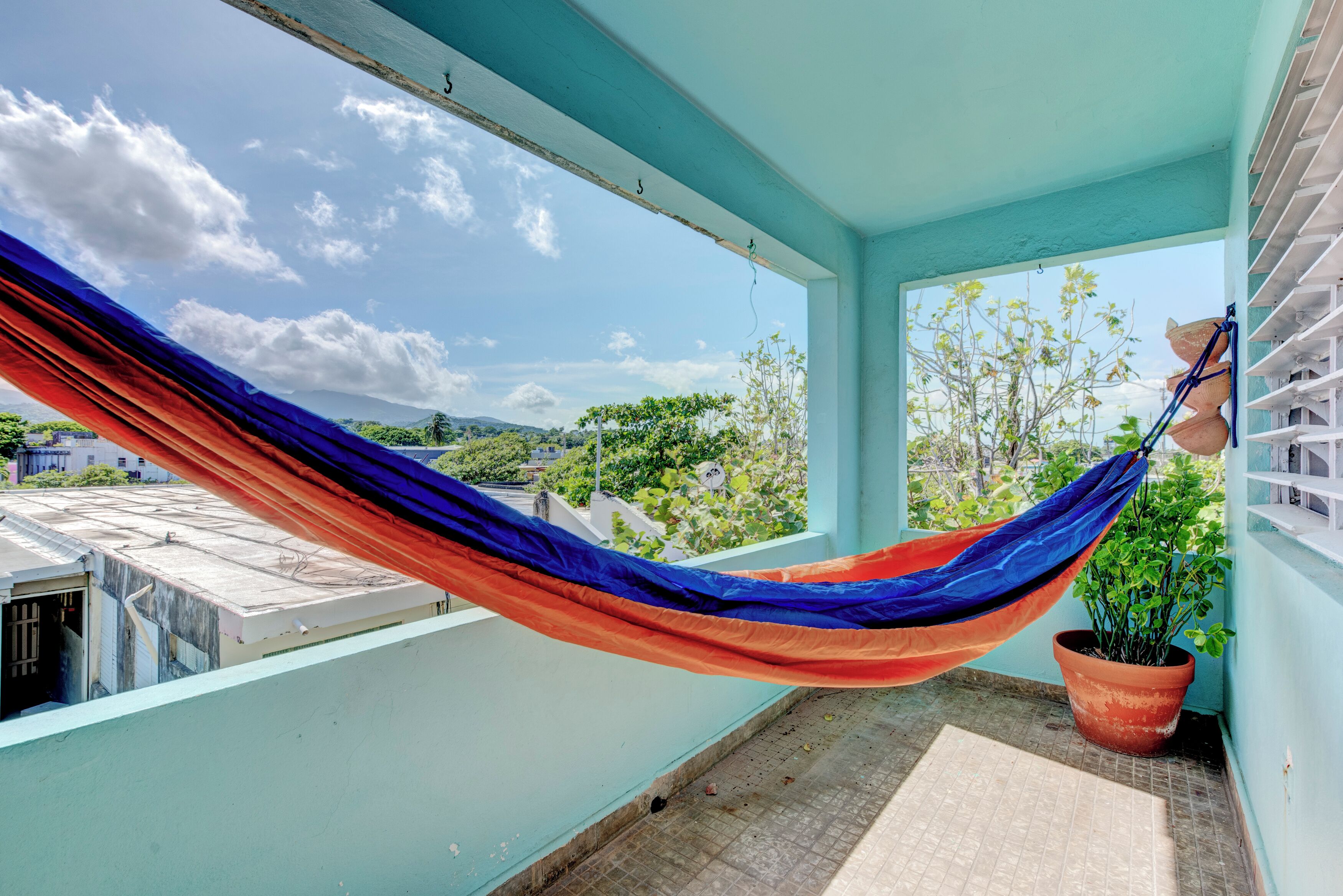 A hammock on a balcony with a potted plant and a view of greenery and buildings.