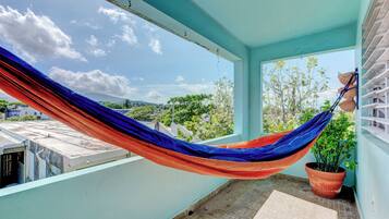 A hammock on a balcony with a potted plant and a view of greenery and buildings.