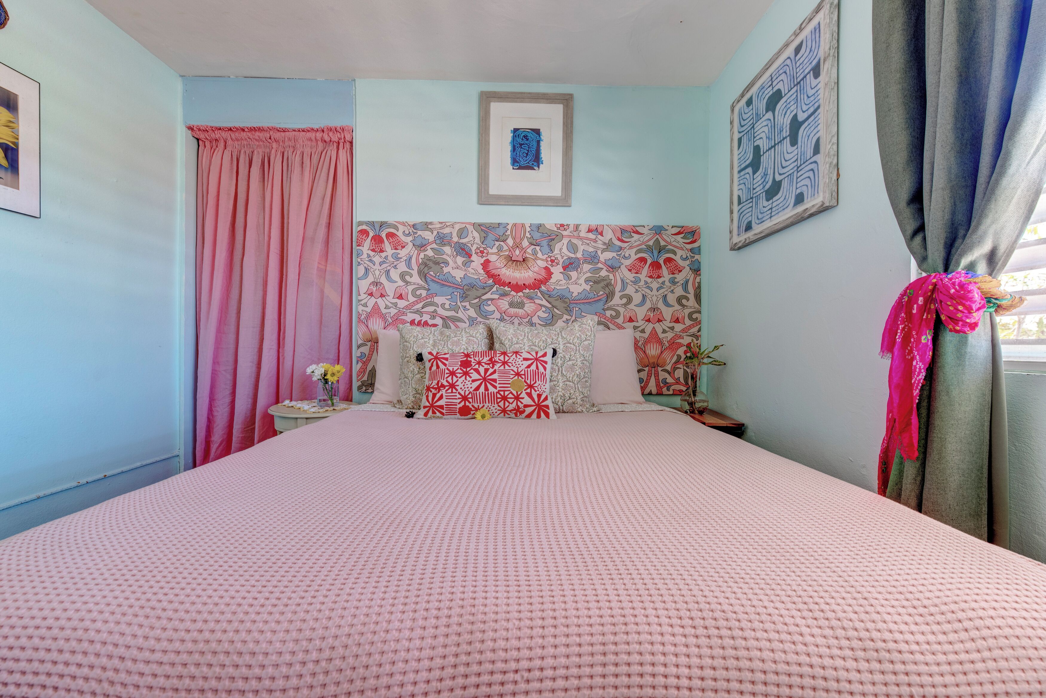 Une chambre à coucher avec un grand lit, une tête de lit à motifs, un rideau rose et un tableau encadré au mur.