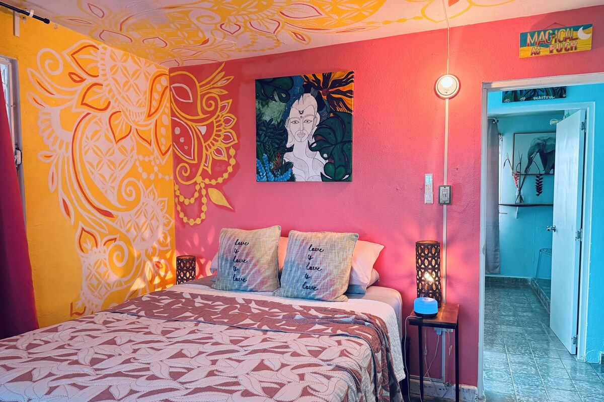 A bedroom with a patterned bedspread, a painting on the wall, and a bedside lamp.