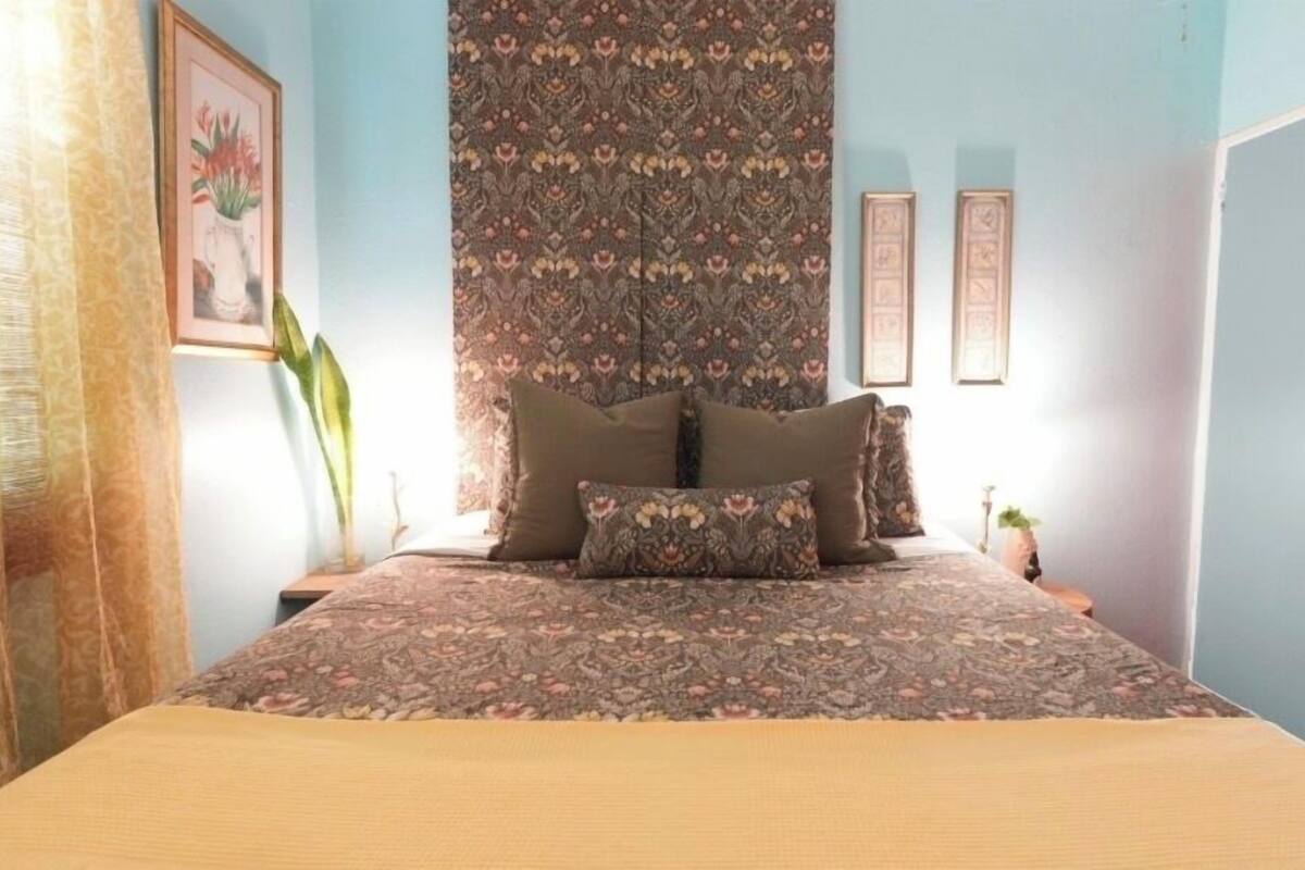 A bedroom with a bed, patterned curtains, a framed picture, and a plant.