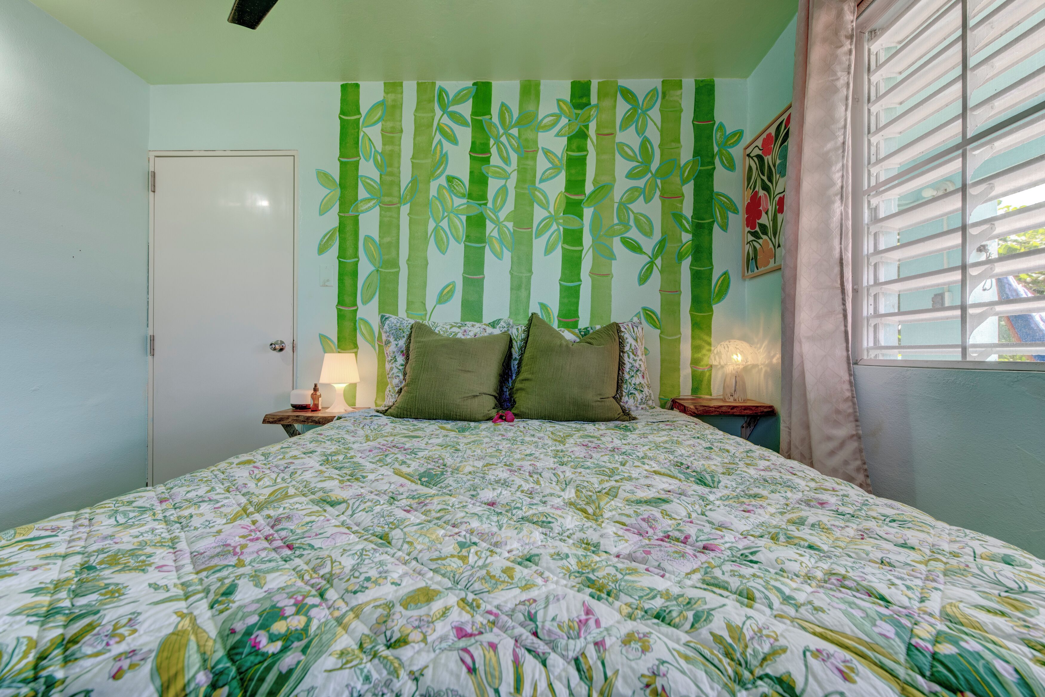 Un dormitorio con colcha estampada, pared con motivos de bambú, una puerta, una mesita de noche con lámpara y una ventana con cortinas.