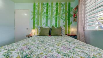 Un dormitorio con colcha estampada, pared con motivos de bambú, una puerta, una mesita de noche con lámpara y una ventana con cortinas.
