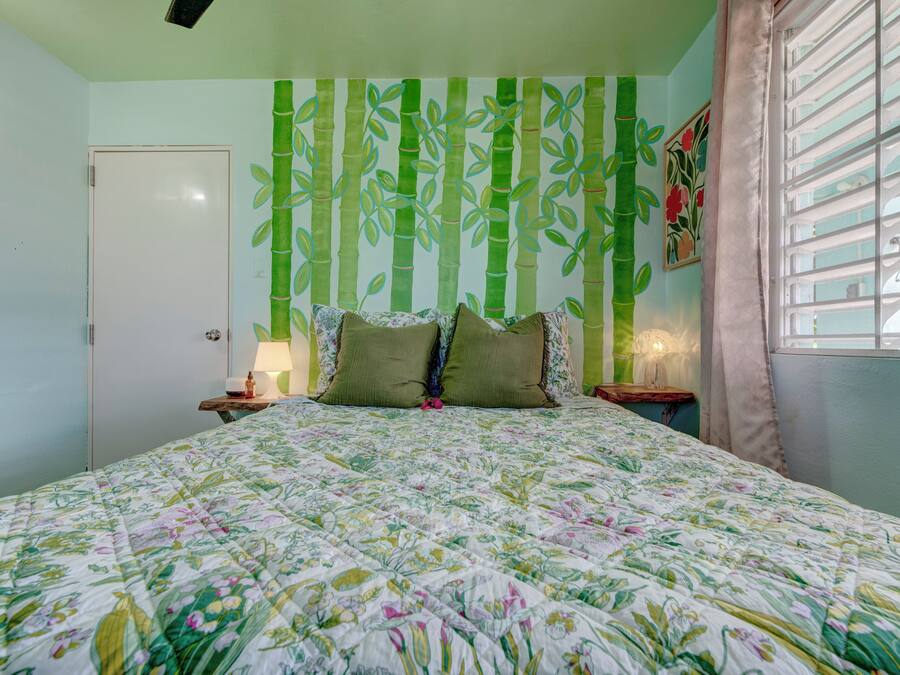 Un dormitorio con colcha estampada, pared con motivos de bambú, una puerta, una mesita de noche con lámpara y una ventana con cortinas.