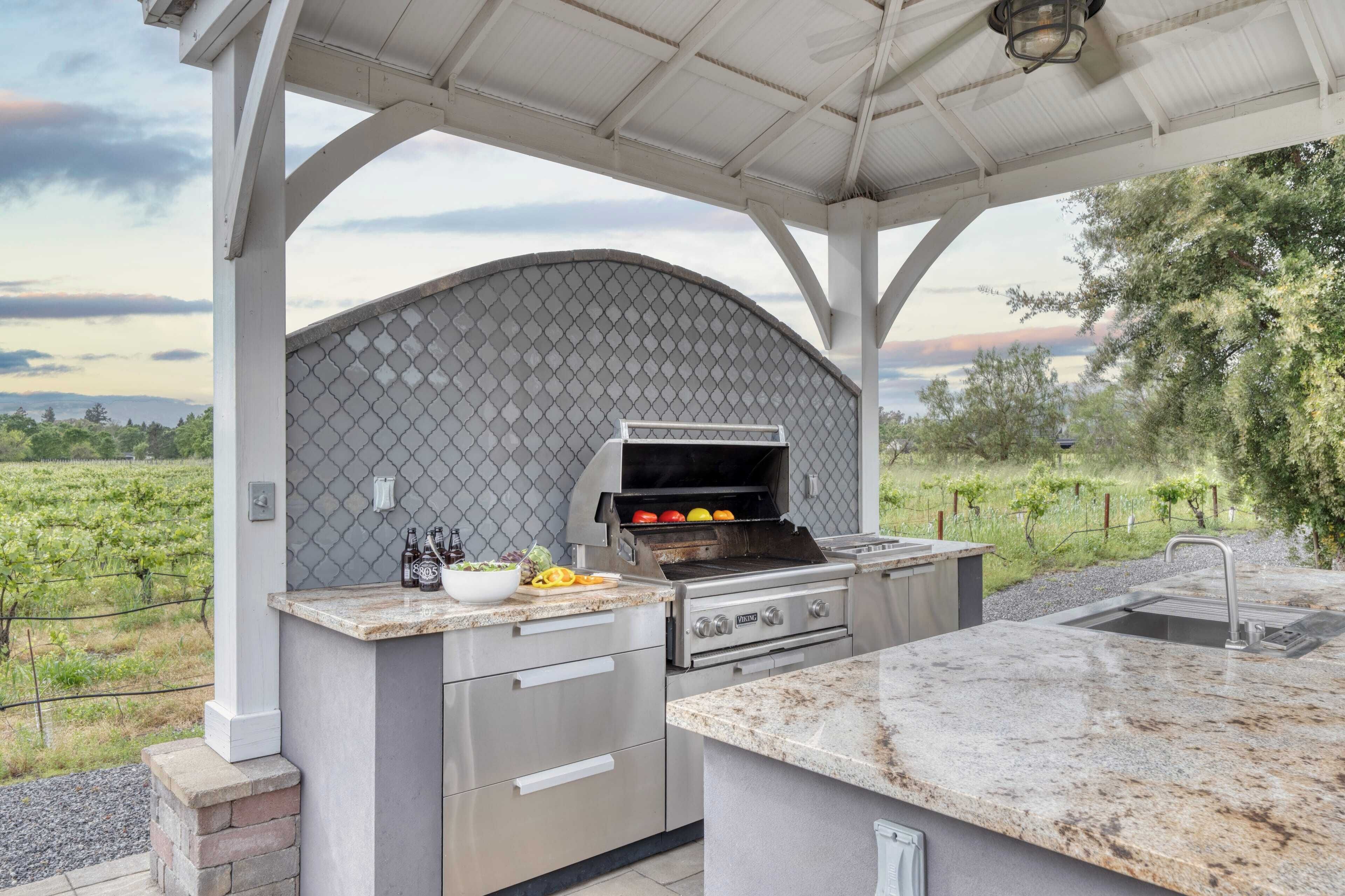 An outdoor kitchen with a grill, a sink, and a countertop with a bowl of fruit and bottles.