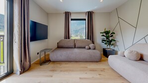 A modern living room with a sofa, a TV mounted on the wall, and a large window with curtains. - Stay Here Living  (Thiersee)