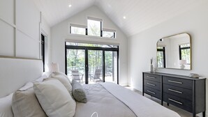Une chambre avec un grand lit, une commode et un miroir. Il y a des fenêtres et une vue sur l’extérieur.