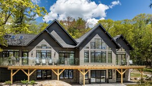 Une maison moderne à deux étages, dotée d’une terrasse en bois, de grandes fenêtres et de fondations en pierre.