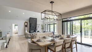 Un espace de repas moderne, avec une grande table dressée pour six personnes, un lustre et une vue sur l’extérieur grâce à de grandes fenêtres.