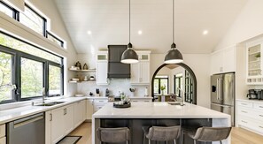 Une cuisine moderne dotée d’un grand îlot central, de luminaires suspendus et offrant une vue sur l’extérieur grâce à de grandes fenêtres.