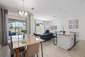 A modern kitchen with a central island, a dining area with a glass table, and a living area with a sofa and a TV.