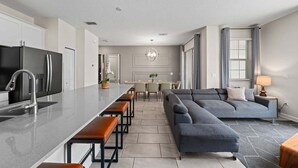 Una cocina moderna con una isla central, un comedor con una araña y una sala de estar con sofá y lámpara.