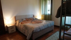 Une chambre à coucher avec un lit, des tables de chevet, une lampe, une fenêtre avec des rideaux et un plancher en bois.