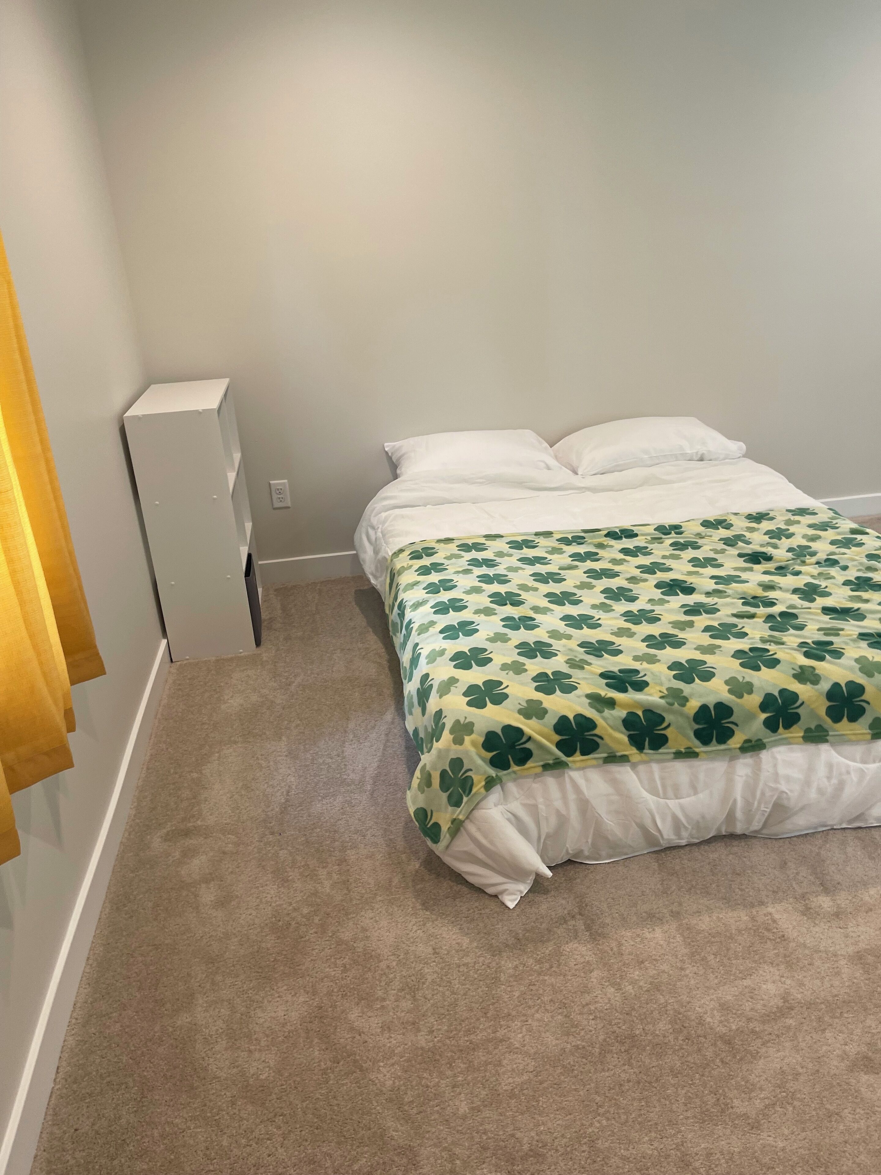 Quarto com cama, almofadas brancas, manta verde e amarela, guarda-roupa branco e piso coberto por carpete.