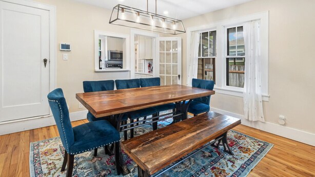 Un coin repas avec une table en bois, des chaises à l’assise rembourrée de couleur bleue, un banc, un lustre et une vue sur la cuisine à travers des portes vitrées.