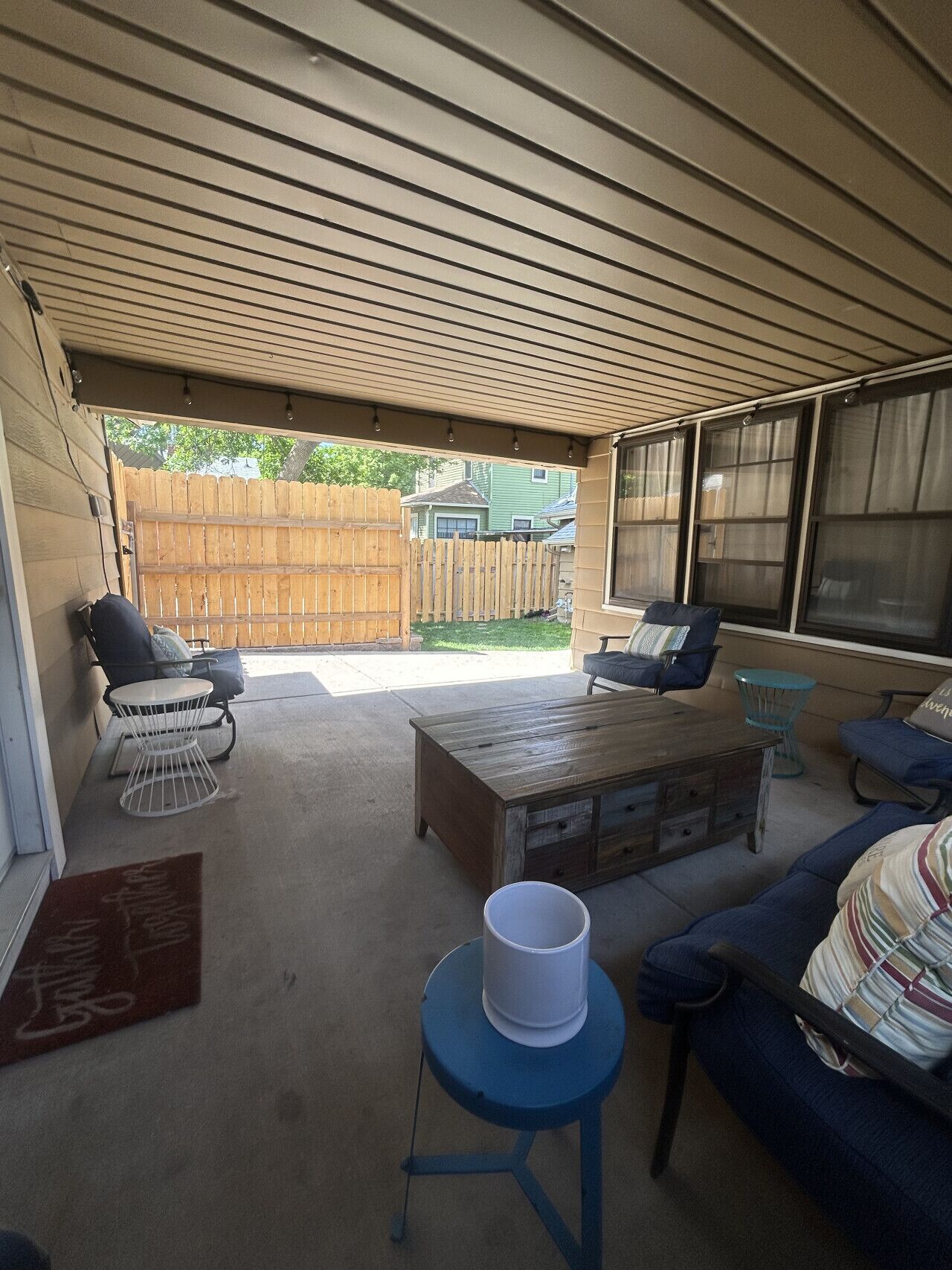 A covered patio area with a wooden table, two chairs, a sofa, and a small round table with a bucket.