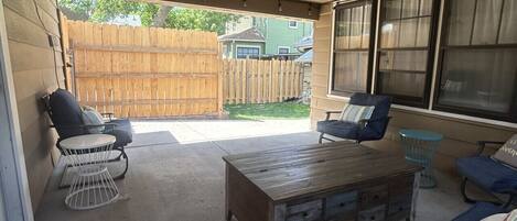 A covered patio area with a wooden table, two chairs, a sofa, and a small round table with a bucket.