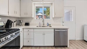 A modern kitchen with white cabinets, a stove, a dishwasher, and a window with a view of a house. - Cozy Salt Lake Getaway - Desk w/ Monitor ! (Salt Lake City)