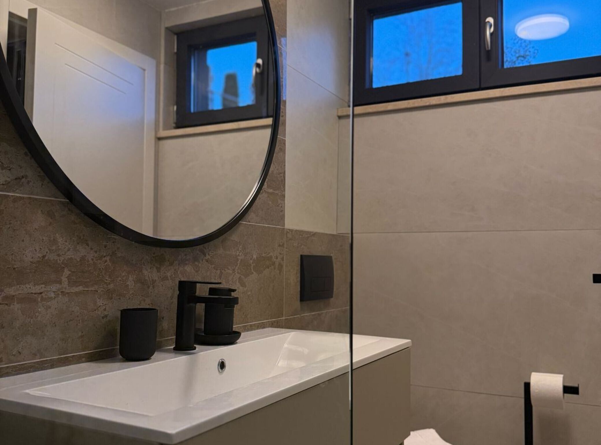 Un baño moderno con un espejo grande, un lavamanos blanco, una cafetera negra y un ventanal con vistas a la ciudad.