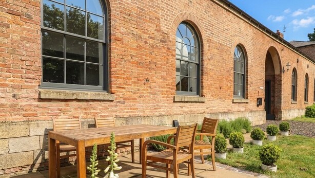 Un bâtiment en briques doté de grandes fenêtres et d’un espace patio avec une table et des chaises en bois.