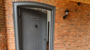 Une porte gris foncé, surmontée d’une enseigne indiquant « WEST STABLE », est encastrée dans un mur de briques, avec une lanterne en métal placée à sa droite.