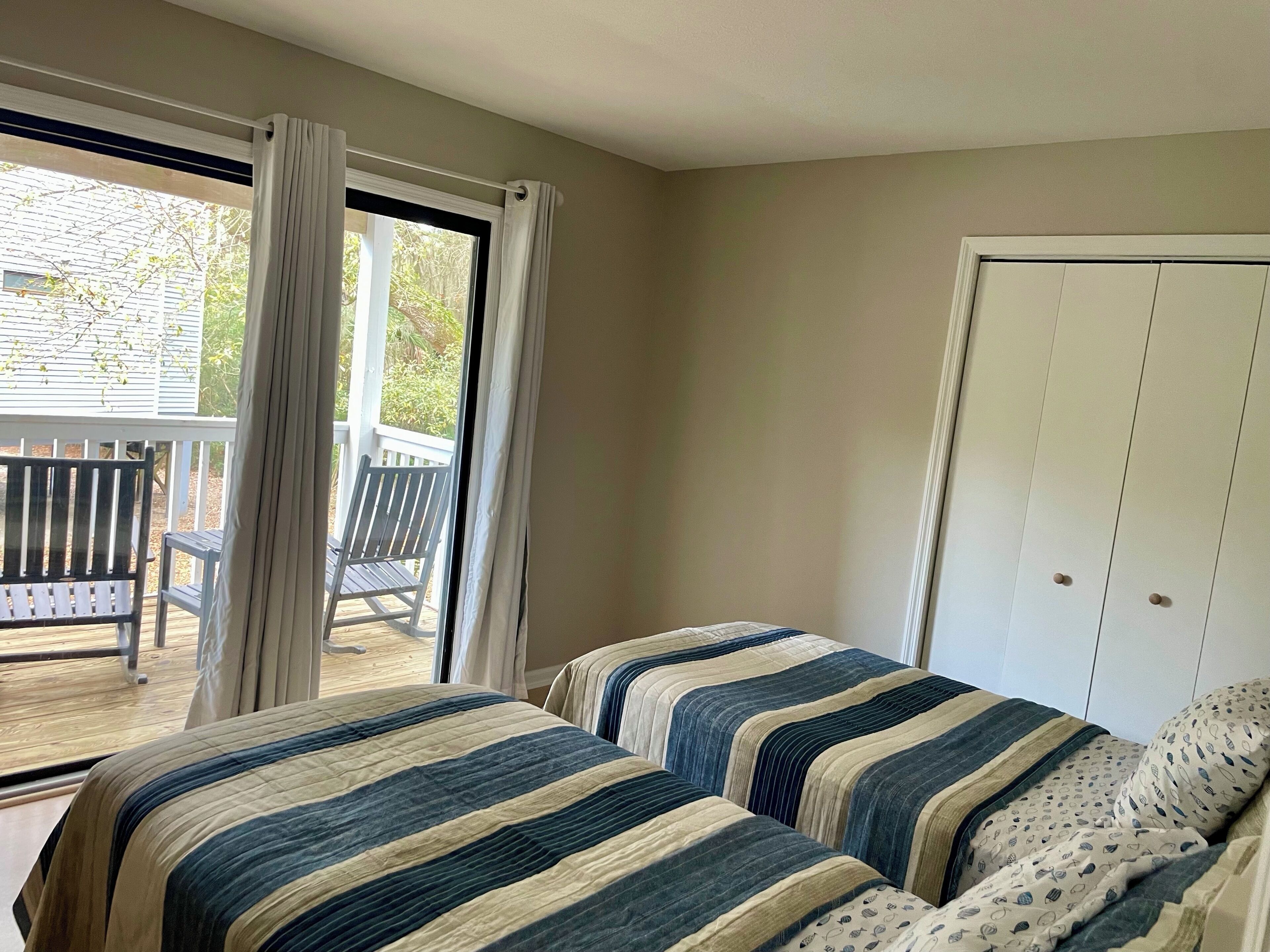 Un dormitorio con dos camas, una ventana con cortinas, vistas a una terraza con sillas y una puerta blanca.