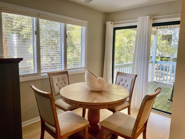 A dining area with a round wooden table and four upholstered chairs. There are three windows with blinds, and a sliding glass door leading to an outdoor deck.