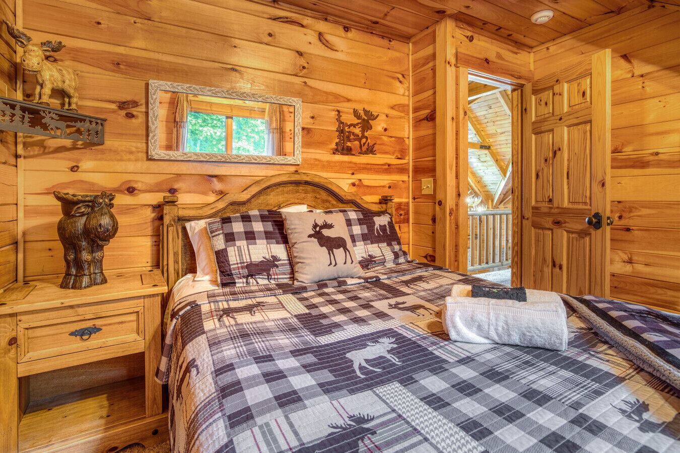 A cozy wooden cabin bedroom with a bed featuring a plaid quilt, a wooden nightstand with a decorative vase, and a window with curtains.