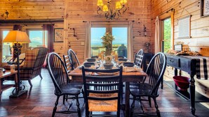 Interno di una baita in legno con un tavolo da pranzo imbandito per sei persone, circondato da sedie, un lampadario e vista panoramica attraverso ampie finestre.