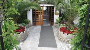 Un chemin menant à un bâtiment doté d’une porte en bois et d’un petit jardin agrémenté de fleurs rouges et de plantes vertes.