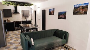 A modern living room with a green sofa, a glass coffee table, and a dining area with a glass table and chairs. There are kitchen cabinets and a stove in the background.