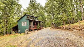 A rustic cabin with a porch, surrounded by dense trees and a gravel driveway.