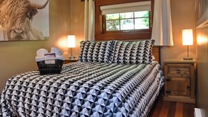 Una cama bien hecha con una colcha de estampado geométrico, una mesita de noche con una lámpara, una pequeña mesita auxiliar de madera y una ventana con cortinas.
